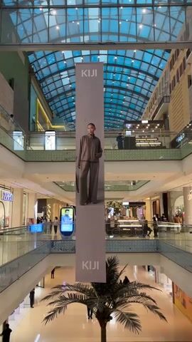 The video shows the interior of a multi-level shopping mall with a glass ceiling. A large banner featuring a model wearing a brown outfit and the brand name "KIJI" is hanging from the ceiling in the center of the frame. Below the banner, a palm tree is visible. Several people are walking around on different levels of the mall, and shops are visible on the sides. The overall lighting suggests it is daytime.