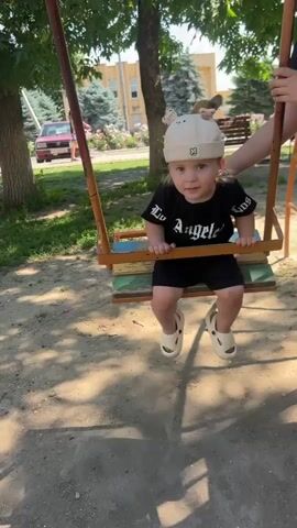 A toddler wearing a white hat with animal ears, a black t-shirt with white text that reads "Los Angeles," and white sandals sits on a weathered swing. The swing is made of wood and metal, with peeling paint on the wooden seat. The toddler's hands grip the wooden front bar of the swing. In the background, a person's arm, likely an adult's, is visible on the right, suggesting they are holding or pushing the swing. The environment appears to be an outdoor playground or park, with trees, grass, and sandy ground. In the further background, several people are visible, including a child and an adult near a building, and a red car parked on a paved area. The lighting suggests it is daytime, likely sunny.