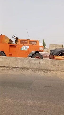 Here's a description of the video:

The video shows two pieces of construction equipment parked on a curb next to a road. The equipment includes a large orange construction roller with "SKY" printed on the side and a smaller, similar roller with "CAT" on the top. Both are parked behind a concrete curb alongside a paved road.  The sky is visible above and behind the equipment, appearing somewhat hazy. Buildings and trees are visible in the background. The camera appears to be moving, as it pans along the curb, focusing on the machinery. There is some traffic noise in the background.