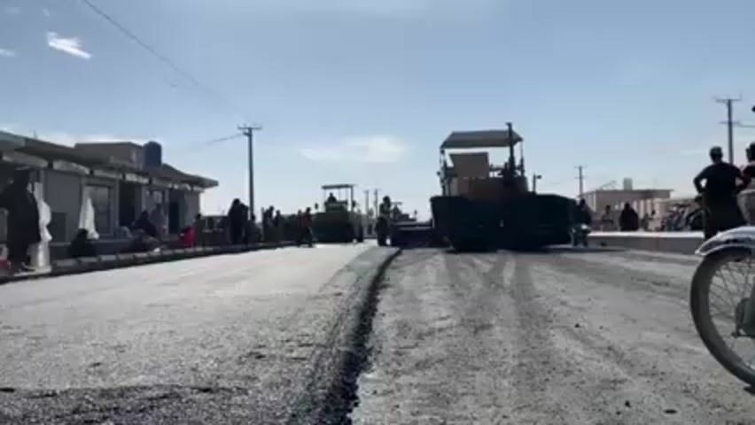 The video shows a road construction scene on what appears to be a sunny day in a populated area. In the foreground, freshly laid asphalt shows a dividing line where the paving has started. A worker in a safety vest walks toward the camera, between the asphalt layer and a paving machine. Another vehicle with a roller is visible behind the paving machine. A group of people, primarily dressed in traditional clothing, sits or stands along the side of the road, observing the work. Buildings line the left side of the road, and power lines stretch across the sky above. A motorcycle is parked to the right of the frame.