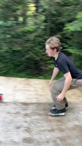 A group of young people are at an outdoor skate park with trees in the background. The video begins with a blurred shot of a person on rollerblades in mid-air, jumping over a red rail. The camera then focuses on the person landing and continuing to skate down a ramp. Other people are watching from the sidelines, some on rollerblades and some standing. The skate park appears to be made of concrete and wood with metal rails. The overall atmosphere seems to be one of recreational activity and possibly a competition or demonstration.