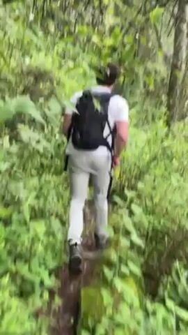 The video shows a person hiking in a lush green forest. The person is wearing a white t-shirt, light-colored pants, and a black backpack. The trail they are walking on is narrow and surrounded by dense vegetation. Other people are ahead on the trail. The video is shot from behind the first person. The forest appears damp, with water droplets on some of the foliage.