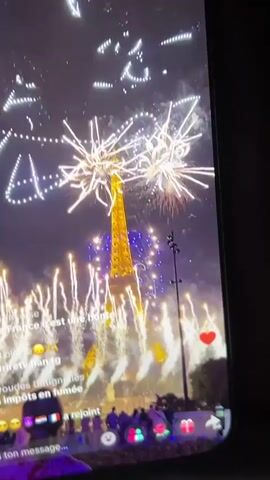 The video shows a nighttime view of fireworks exploding in the sky above the Eiffel Tower. The Eiffel Tower is illuminated and appears golden, with blue lights also visible on its structure. The fireworks are varied in shape and color, with some appearing as bursts of light and others forming patterns in the sky. There are people gathered below the tower, though they are not clearly visible. A live stream interface is visible at the bottom of the screen, displaying comments from viewers. The video appears to be a screen recording of a live social media broadcast.