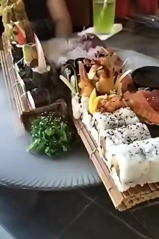 A platter of sushi is presented on a bamboo mat, with a dramatic display of dry ice creating a smoky effect. The sushi includes various rolls with rice and seaweed, topped with ingredients like sesame seeds, avocado, and possibly seafood such as shrimp and salmon. A small side of green seaweed salad, sprinkled with sesame seeds, is also visible. The platter is placed on a large, white, decorative plate. In the background, a person's hand and arm are visible, suggesting someone is about to partake in the meal. A bright green drink in a glass with a straw sits on a table further in the background, alongside a red object that might be another drink or container. The scene appears to be indoors, likely in a restaurant, given the presentation and the presence of other diners implied by the background elements. The overall impression is one of a carefully prepared and presented meal, with the dry ice adding an element of theatricality.