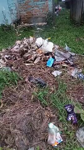 A pile of trash is burning in a grassy yard near a building. A person in a headscarf is seen walking away from the pile and into the frame. The trash consists of leaves, branches, plastic bags, and other refuse. There are also some wrappers on the ground near the pile.