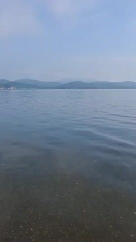 A person stands in shallow water near the shore of a calm lake. The sky is overcast with a hazy blue color. Gentle waves ripple across the surface of the water, and the lakebed is visible through the clear water. In the distance, mountains rise on the horizon. The person is wearing blue shorts and appears to be interacting with the water.