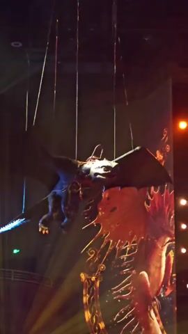 A dragon puppet, suspended by wires, appears to fly in front of a large screen displaying an image of a dragon. The puppet is dark-colored with large wings. The screen shows a detailed image of a red and gold dragon with intricate patterns. The background is dark, with some stage lighting visible, including yellow and white spotlights. The overall impression is of a theatrical performance or a demonstration of special effects.