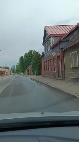 A first-person perspective video shows a car driving on a wet paved road in a village or small town. The weather appears overcast. The road is lined with buildings on both sides. On the right, there are several brick buildings, one of which has a red roof and decorative architectural elements. Trees and some vegetation are visible along the road. On the left side of the road, there are more buildings and trees. The camera is positioned inside the car, showing part of the dashboard at the bottom of the frame. The video is brief, capturing a few seconds of the drive.
