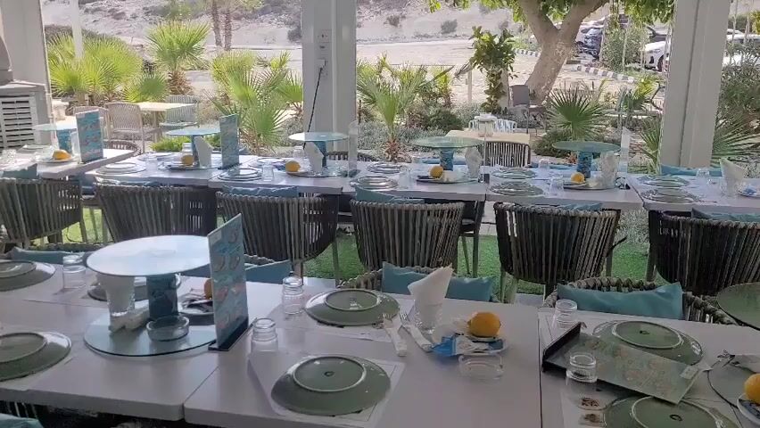 The video shows an outdoor dining area of a restaurant. The tables are set with white tablecloths, plates, glasses, and lemons as decoration. The chairs are woven with a grey rope and have light blue cushions. In the background, there are palm trees and other greenery, as well as a few parked cars. The overall impression is a bright and airy space, possibly by the sea or in a tropical location. The tables are arranged in rows, suggesting a setup for a large group or event.