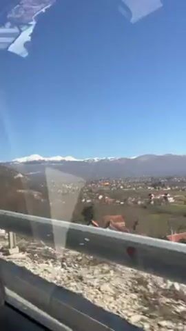 The video shows a view from inside a moving vehicle.  A metal guardrail is in the foreground. Through the window, a landscape unfolds featuring a town nestled in a valley with mountains in the distance. The mountain peaks are capped with snow, contrasting with the clear blue sky above. The town has a mix of building styles, and the surrounding terrain transitions from sparse vegetation near the road to denser patches further out.  The vehicle is moving at a moderate pace, and the camera focuses on the scenery passing by.