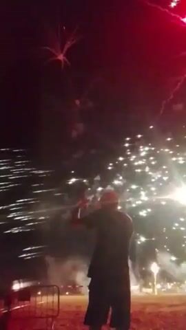 A fireworks display is captured, with a person silhouetted in the foreground, facing away from the camera and towards the sky. The person is wearing a dark coat and appears to be standing on a raised platform or surface. The sky is filled with bursts of red and white fireworks, creating a vibrant and dynamic spectacle. The fireworks are in various stages of explosion and descent, with some appearing as bright sparks and others as large, blooming patterns of light. The overall impression is of a festive and celebratory event, likely taking place at night.