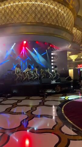 A group of dancers wearing black ski masks and revealing outfits perform on a stage. The stage is lit with red and blue spotlights, creating dramatic effects. The dancers are in various poses, some bending over and others striking more upright stances. The background appears to be a dimly lit casino or entertainment venue, with ornate gold and black patterned flooring in the foreground. There are speakers and lighting equipment visible on the stage. A sign with Cyrillic lettering is present on the stage.