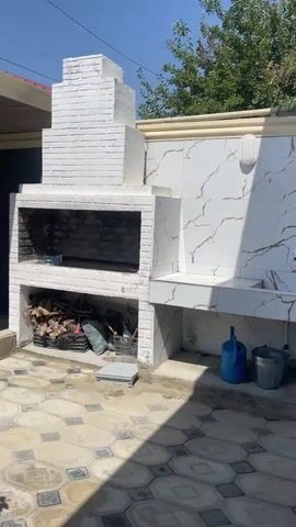 This video shows an outdoor patio area with a brick barbecue grill and a covered seating area. The grill is white and has a chimney. It is stocked with firewood and other items in the lower compartments. To the right of the grill is a countertop with a sink. The patio floor is tiled with a geometric pattern. The covered seating area has a table and chairs. There are rose bushes in the foreground and to the left of the seating area. The sky is clear and blue.