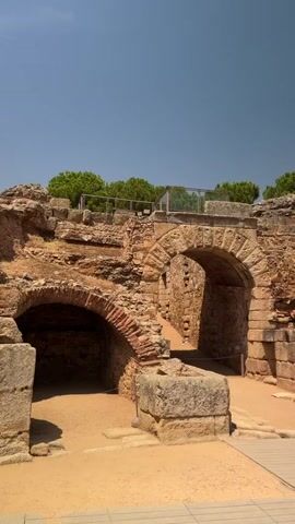 The video pans across the ruins of an ancient stone amphitheater on a sunny day. The structures are made of rough-hewn stone and brick, with arched doorways and crumbling walls. In the foreground, a sandy area leads to the ruins, and a modern wooden walkway is visible. A few tourists, including a man and a woman, are standing and looking at the ruins. One person is taking a photograph. The sky is clear blue, and green trees are visible on the top of the amphitheater. The scene evokes a sense of history and the passage of time.