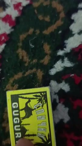 A hand strikes a match against a matchbox to light it, then holds the lit match. The matchbox is yellow, and has an image of a camel and palm trees on it, as well as Cyrillic text. The background is a patterned rug.