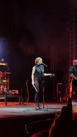 A woman is performing on stage in front of a crowd at an outdoor concert venue in Kragujevac, Serbia. She is wearing a black sleeveless top, sparkly shorts, black leggings, and platform boots. She is holding a microphone and a drumstick. There is smoke and stage lighting, with orange and blue lights illuminating the stage. A drummer and a guitarist are visible behind her. The concert appears to be taking place at night. The audience is partially visible in the foreground, with some holding up their phones to record the performance. The woman's expression suggests she is actively engaged in her performance, possibly singing or speaking to the crowd.