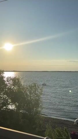 The video shows a serene lake scene at sunset. The sun is low on the horizon, casting a warm glow and a sunbeam across the water. In the middle distance, three figures are visible on a small boat or raft. They appear to be enjoying the water, with one person potentially standing. The shoreline is visible in the foreground, with trees and some vegetation. The water is calm, with gentle ripples. The sky is clear with a few wispy clouds. A white car is partially visible on the right side of the frame.