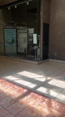 This video shows a walk-through of an indoor space with tiled floors and glass display windows.  Sunlight is streaming in from the right, casting shadows on the floor. In the display window on the right, there is a poster for the Clinton County Historical Association and another poster about the Battle of Valcour Island. There are also some artifacts visible in the display. To the left of the historical display, there is another glass-fronted display case with items inside. In the background, a yellow vehicle and part of another display can be seen.