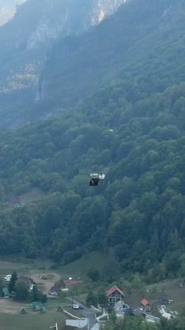 This is a video of a cable car moving across a forested mountain landscape. The cable car is carrying passengers and appears to be in motion. The surrounding environment is dominated by lush green trees covering the slopes of the mountain. In the foreground, there are some buildings and a road, suggesting a small settlement or village at the base of the mountain. The sky is hazy, and the lighting suggests it is either early morning or late afternoon. The cable car is the primary focus of the video, as it traverses the vast expanse of the forest.