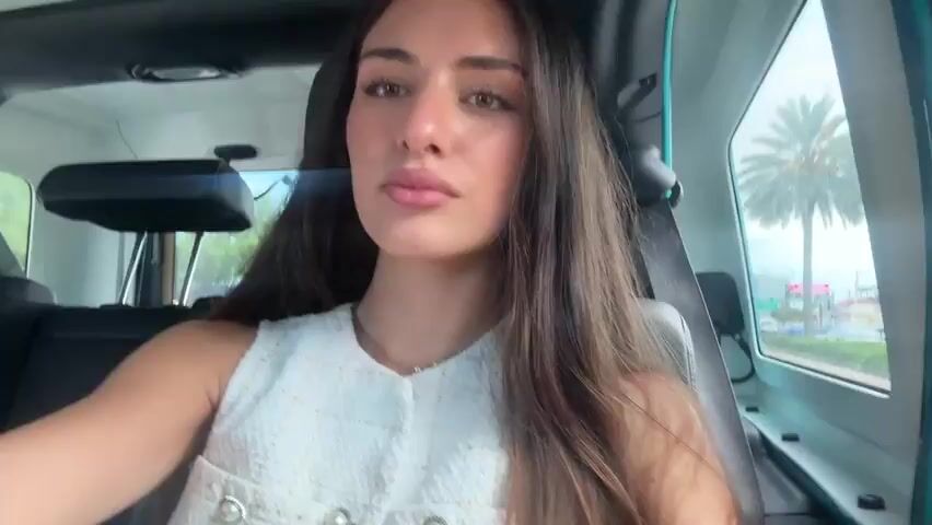 A woman with long dark hair and wearing a white sleeveless top with pearl buttons is sitting in the passenger seat of a vehicle. She is looking at the camera, puckering her lips, and winking. The interior of the vehicle is visible, including the headrest of the driver's seat and a portion of the dashboard. Outside the window, palm trees and a cloudy sky are visible. The lighting suggests it is daytime, possibly overcast or raining.