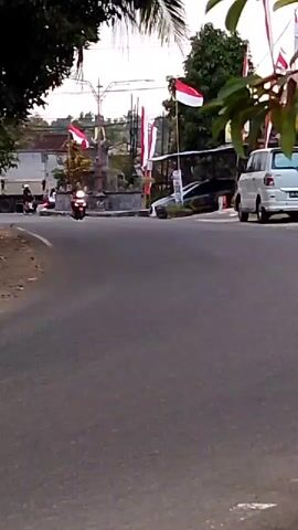A motorcycle with a rider is moving towards the camera on a paved road. Indonesian flags are visible in the background, along with trees and buildings. A white van and a black pickup truck are parked on the side of the road. The sky is overcast.