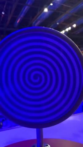 A close-up shot focuses on a large, circular object with a blue and black spiral pattern. The object appears to be a prop or an exhibit, possibly in an entertainment venue or museum, indicated by the blurred background lights suggestive of a ceiling with overhead lighting fixtures. The spiral on the object is concentric, with the lines appearing to draw the viewer's eye inward. The lighting in the scene is predominantly blue, casting a glow on the surface and surrounding area. The object is mounted on a metallic pole, which is partially visible at the bottom of the frame. The overall impression is one of a visually engaging and perhaps disorienting design.