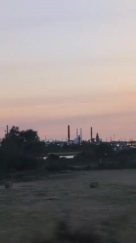The video begins with a wide shot of a field with two hay bales visible in the foreground. In the middle ground, there is a body of water and a line of trees. Beyond the trees, a distant industrial complex with numerous tall smokestacks and buildings is silhouetted against a pale pink and white sky. The sky suggests either dawn or dusk. The camera then pans slightly to the left, revealing more of the trees in the foreground and a more expansive view of the industrial area. The lighting is dim, and the overall scene is quiet and still.