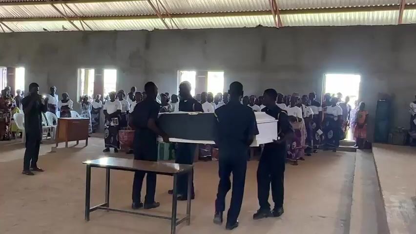 A group of pallbearers in dark uniforms carry a dark casket inside a sparsely furnished building. They are walking towards a table placed in the foreground of the image. A crowd of people dressed in white shirts and colorful patterned skirts are gathered on both sides of the room, watching the procession. Some individuals in the crowd are holding up cell phones, likely to record the event. The building appears to be a community hall or a church, with bare concrete walls and a corrugated metal roof. Natural light streams in through open doorways and windows. The floor is dirt.