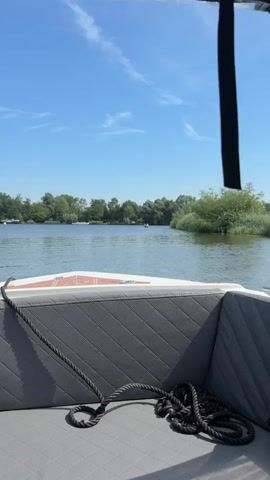 A first-person perspective video of a boat moving on a body of water. The foreground shows a grey quilted boat seat with a black rope coiled on it. The boat is moving forward, indicated by the slight blur of the water. In the middle ground, the water is calm with a few ripples. On either side of the waterway are lush green trees and reeds. In the distance, there are more trees and some buildings are visible on the left bank. Two people are visible in a small boat on the water in the distance, further up the waterway. The sky is a clear blue with a few wispy white clouds. The sun is shining brightly, casting shadows on the boat seat. The overall impression is a peaceful, sunny day on the water.