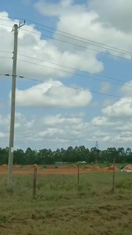 Here is a concise description of the video:

The video shows a rural landscape scene captured from a moving vehicle. In the foreground, there's a grassy field with piles of reddish-brown material, possibly sand or soil. A simple wooden fence runs along part of the field. In the background, there is a line of trees under a sky with scattered white clouds. Utility poles and power lines stretch across the sky. As the video progresses, a few small structures and houses become visible amidst the trees.
