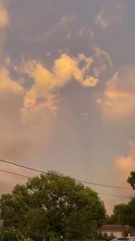 The video shows a rainbow in a cloudy sky at sunset. The sky is a mix of pink, orange, and blue hues, with fluffy clouds scattered throughout. The rainbow is a vibrant arc of color, stretching across the sky. Below the sky, there are trees and houses, as well as power lines. The overall scene is peaceful and serene.