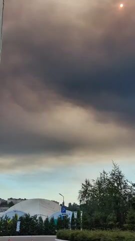 The video shows a sky with dark clouds and a bright sun in the upper right corner. In the lower part of the frame, there are trees and a white structure, possibly a building. A blue and white sign with a letter "P" on it is visible.