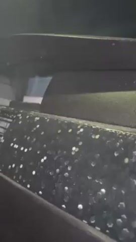 The video shows the interior of a car at night. The camera starts focused on the dashboard, which has a black, speckled pattern, and then pans upward to show the car's display screen, air vents, and a small red figure hanging from the rearview mirror. The camera continues to pan up and to the left, revealing the steering wheel, speedometer, and tachometer, which have red illumination. An air freshener hangs from the rearview mirror. The view through the windshield is dark, with a few lights visible in the distance.