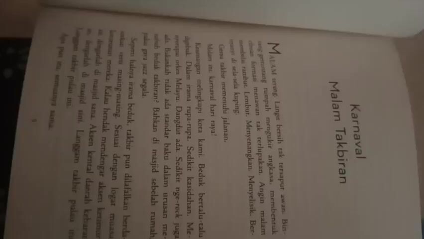 The video shows a person's hands holding an open book. The book has a title on the right page that reads "Karnaval Malam Takbiran." The text on the left page is in Indonesian. The video is blurry and the image is rotated, making it difficult to read the text clearly. The lighting is dim, and the background is out of focus. The person's hands are visible, with fingers turning the pages of the book.