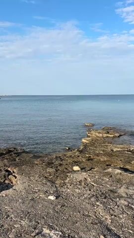 A vertical video captures a view of the ocean and a rocky shoreline under a blue sky with scattered white clouds. The foreground shows a rough, textured surface of rocks and dirt, with a single white object, possibly litter, on the ground. Beyond this, the clear, calm water of the ocean stretches towards the horizon. There are a few scattered rocks or small islands emerging from the water in the middle ground. The sky is a bright, clear blue with wispy white clouds. The lighting suggests it is daytime. No people are visible in the frame, nor are any boats or other significant man-made objects in the water or on the shore.