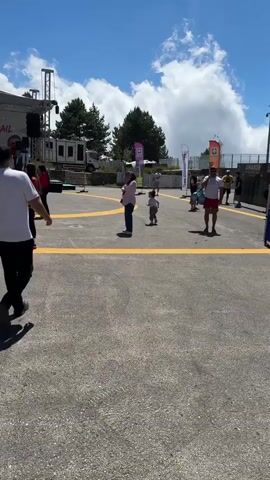 This media captures an outdoor event, likely a race finish line, on a sunny day. In the foreground, a paved area with yellow lines marks a pathway. Several people are visible, some walking away from the camera and others facing towards a stage area. 

The backdrop features a stage with a large banner that reads "AMANOSTRAIL" and "HOŞ GELDİNİZ!", which translates to "Welcome!". A group of people, some wearing red shirts and others blue, are lined up behind a finishing tape stretched across the area in front of the stage. There are also flags of Turkey visible. The setting appears to be a natural landscape with a dense forest of pine trees and a clear blue sky with scattered clouds. A tall telecommunications tower can be seen in the distance, as well as tents and other structures related to an event. 

The overall mood seems celebratory and active, with participants and spectators gathered for the trail event. The bright sunlight and clear sky suggest it is daytime. The presence of the banner and the finishing tape strongly indicates a sporting event, likely a trail race given the "TRAIL" text.