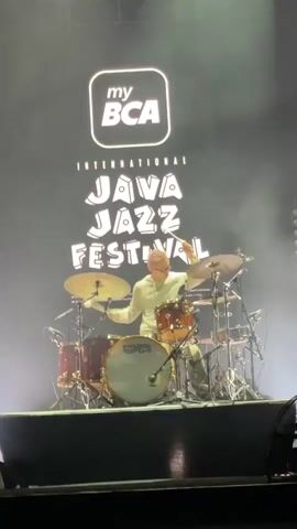 A drummer plays his kit on stage at the International Java Jazz Festival. The backdrop features the festival's logo and the sponsor, my BCA. The drummer, wearing a white hoodie, is surrounded by his drum set, which has a "Wave to Earth" logo on the bass drum. He is illuminated by stage lights, with a slight haze in the air. The stage is dark, and some audience members are visible at the bottom of the frame.