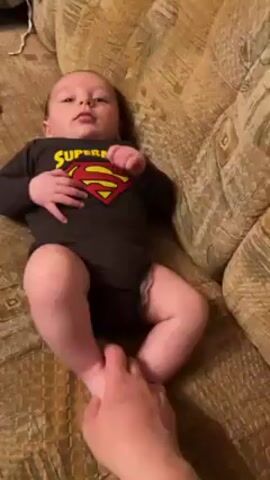 A young, light-skinned infant wearing a black Superman onesie lies on a gold textured sofa. An adult hand holds the infant's feet and moves them in a tickling motion. The infant looks around and makes sounds.