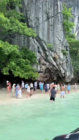 The video shows a group of people standing on a sandy beach with crystal clear turquoise water lapping at their feet. Towering limestone cliffs covered in lush green foliage rise dramatically behind them. The people, dressed in a mix of casual beachwear and modest attire, appear to be tourists, some are gathered in small groups, while others are standing individually. The water is shallow and calm, suggesting a sheltered cove or bay. The overall atmosphere appears relaxed and serene, with the natural beauty of the environment being the focal point.
