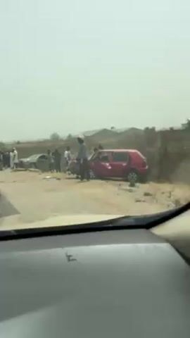 From the perspective of a driver inside a vehicle, the video shows a car accident scene on a dirt road. Two vehicles are visibly damaged, one is red and the other is silver. Several individuals are standing near the wreckage, observing the scene. Some appear to be wearing casual clothing while others have on what looks like work uniforms or vests. The environment is dusty with sparse vegetation and some distant buildings. The sky is overcast, casting a diffused light over the scene.