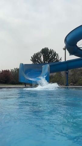 A boy in a blue long-sleeved rash guard and red shorts emerges from the bottom of a blue waterslide into a blue swimming pool. Water splashes as he enters the pool. The waterslide has a spiral design at the top. Behind the pool and waterslide, the sky is overcast, and a tall, branching cactus-like plant is visible. Other trees and landscaping are present.