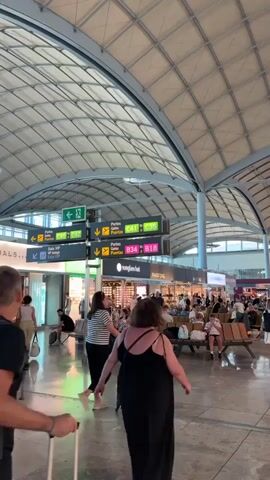 The video shows a busy airport terminal. Several people are walking, some pulling luggage, others with backpacks.  Many people are seated on benches in a central waiting area.  Above, there are signs in multiple languages directing travelers to gates C41 to C35 and B34 to B18.  Shops like Sunglass Hut and Parfois are visible in the background. The airport features a high, arched ceiling with a grid-like pattern. The floor is made of glossy tiles.