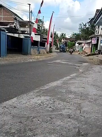 A view of a road with several buildings on the left and right. Indonesian flags are visible on the left. Several motorbikes and a truck are driving on the road. The first motorbike has a rider facing left, the second motorbike has a rider facing right, and the third motorbike has a rider facing right. The truck is dark colored and covered with a blue tarp. The sky is cloudy.