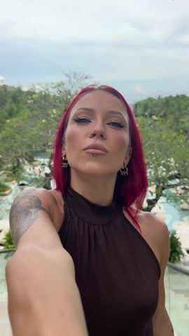 A woman with dark red hair, tattoos on her arm, and multiple piercings stands in front of a water feature taking a selfie video. She is wearing a brown sleeveless top, and gold-colored earrings. In the background are trees and a turquoise waterway. Another person then walks up the stairs behind the woman, disrupting the selfie. He is wearing a tank top and shorts and carrying a jacket. He walks towards the top of the frame.