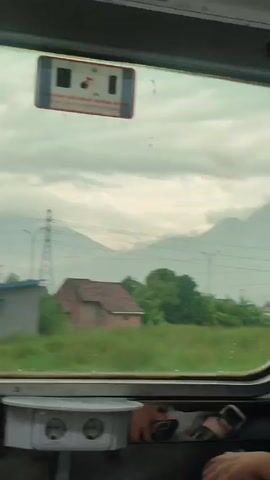 Here is a concise description of the video:

The video shows a view from inside a moving train. The camera is focused on the window, through which various houses and buildings are seen passing by. In the background, a mountain is visible under an overcast sky. There are signs on the window in Indonesian, and an electrical outlet below. The overall environment seems to be a suburban or rural area.