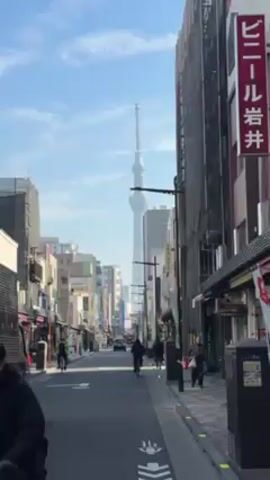 The camera, moving along a narrow city street under a bright sky, frames the Tokyo Skytree at the street's end.  Buildings line both sides of the street, with pedestrians, cyclists, and cars passing.  A commercial vehicle is parked at the street’s edge in the foreground, and signage in Japanese is visible on the buildings. Scaffolding or netting covers the facade of one building on the right, contrasting with the clear view of the Skytree.
