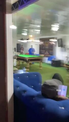 The video shows an indoor pool hall with several pool tables. In the foreground, a person wearing a gray cap and black clothing is reclining on a blue couch, looking at a smartphone. In the background, two men are playing pool at a table. One man, wearing a blue shirt, is standing over the table with a cue stick. The other man, wearing a red shirt, is standing to the side. Another pool table is visible to the right with a person sitting at it. The room has green carpeting and a white ceiling with overhead lights. A poster with text and images is hanging on the back wall. The lighting is artificial.