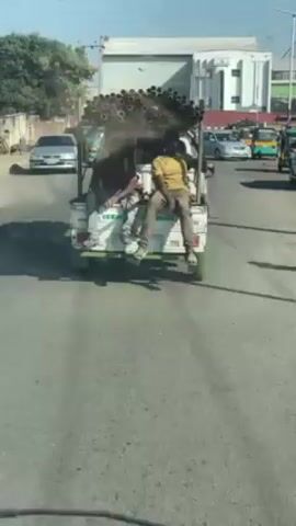 From inside a vehicle, the video shows a small, three-wheeled truck driving on a city street. The truck is overloaded with a large bundle of metal pipes on its roof. Two men are sitting on the tailgate of the truck, casually dangling their legs. Other vehicles, including cars and motorized rickshaws, are also present on the road. The environment appears to be an urban area with buildings lining the street. The video seems to have been taken during daylight.