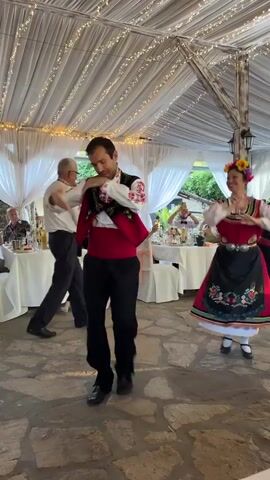 This video shows a lively scene taking place outdoors under a draped canopy adorned with fairy lights. The flooring is paved with uneven, large stones. In the foreground, a man in traditional Bulgarian attire, including a red vest with embroidered detailing and a white shirt with patterned sleeves, is energetically dancing. He is performing a traditional folk dance with synchronized arm and leg movements.

To his right, a woman also dressed in traditional Bulgarian costume, featuring a red dress, a decorated apron, and flowers in her hair, is dancing with a bright smile. Her movements appear to be synchronized with the man's. In the background, other people are seated at tables, some watching the dancers. Another man in similar attire is visible in the mid-ground, also dancing. The presence of tables with white tablecloths suggests a celebratory event, possibly a wedding or a cultural festival. The setting appears to be a garden or an outdoor dining area, with hints of greenery visible beyond the canopy. The lighting suggests it could be late afternoon or early evening, given the fairy lights. The overall mood is joyous and festive, with the dancers clearly enjoying themselves and engaging the audience. There is no visible text in the video.