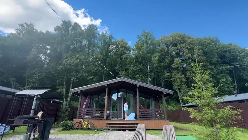 A low-angle shot shows a wooden cabin with large glass windows and a porch. A person in a white shirt and blue shorts stands at the entrance of the cabin. To the left of the cabin, there is a grill and a yellow bicycle parked in front of it. In the background, a dense forest with tall trees is visible under a bright blue sky with scattered clouds. Additional small, dark-colored structures with pitched roofs are situated on either side of the cabin. The ground in front of the cabin is covered with gravel, and there are wooden planks leading up to the entrance. The overall setting appears to be a resort or a glamping site.